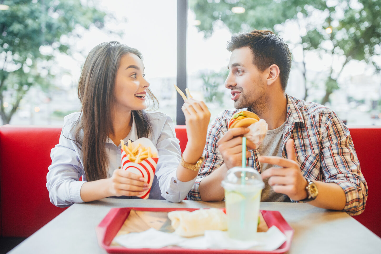 Young beautiful friends have fun talking, looking at each other and eating tasty burger and fries at the window at the fast food restaurant. 2023/08/AdobeStock_231388919.jpeg 