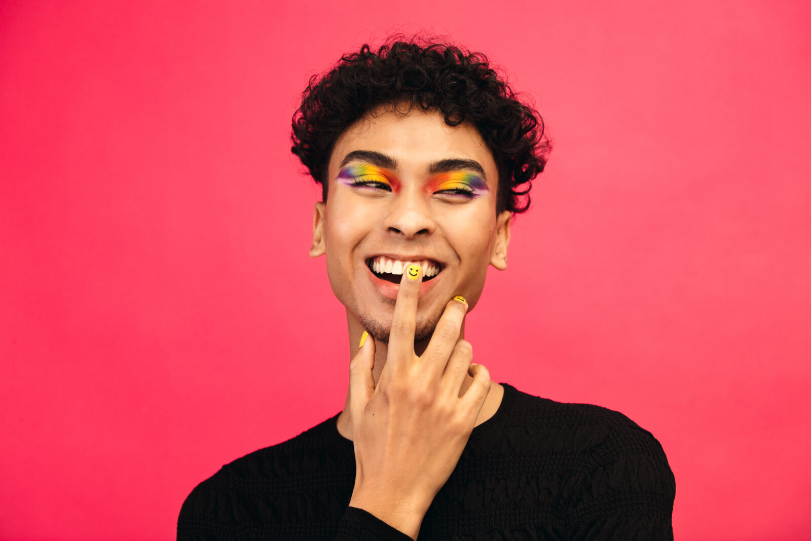 Happy young man wearing rainbow colored eye shadow and smile face nail paint. Gay male smiling with hand on face against red background. 2023/11/AdobeStock_382407018.jpeg 