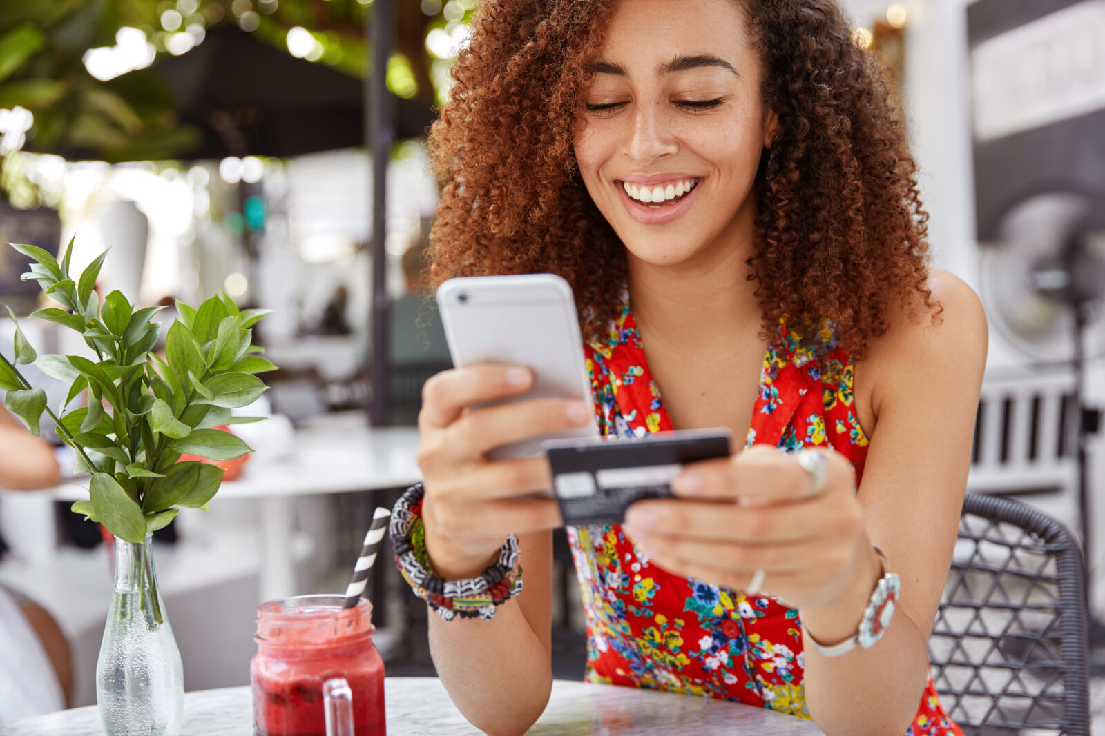 Beautiful dark skinned young female with cheerful expression, holds smart phone and credit card, banks online or makes shopping while sits against cafe interior. Payment and leisure concept. 2024/09/AdobeStock_198775702.jpeg 