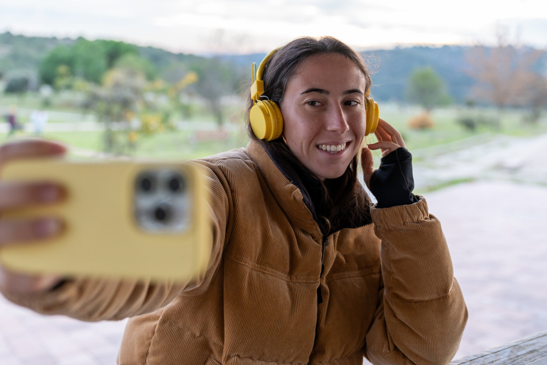 Woman smiling, taking a selfie with a smartphone, listening to music on yellow headphones in a natural park